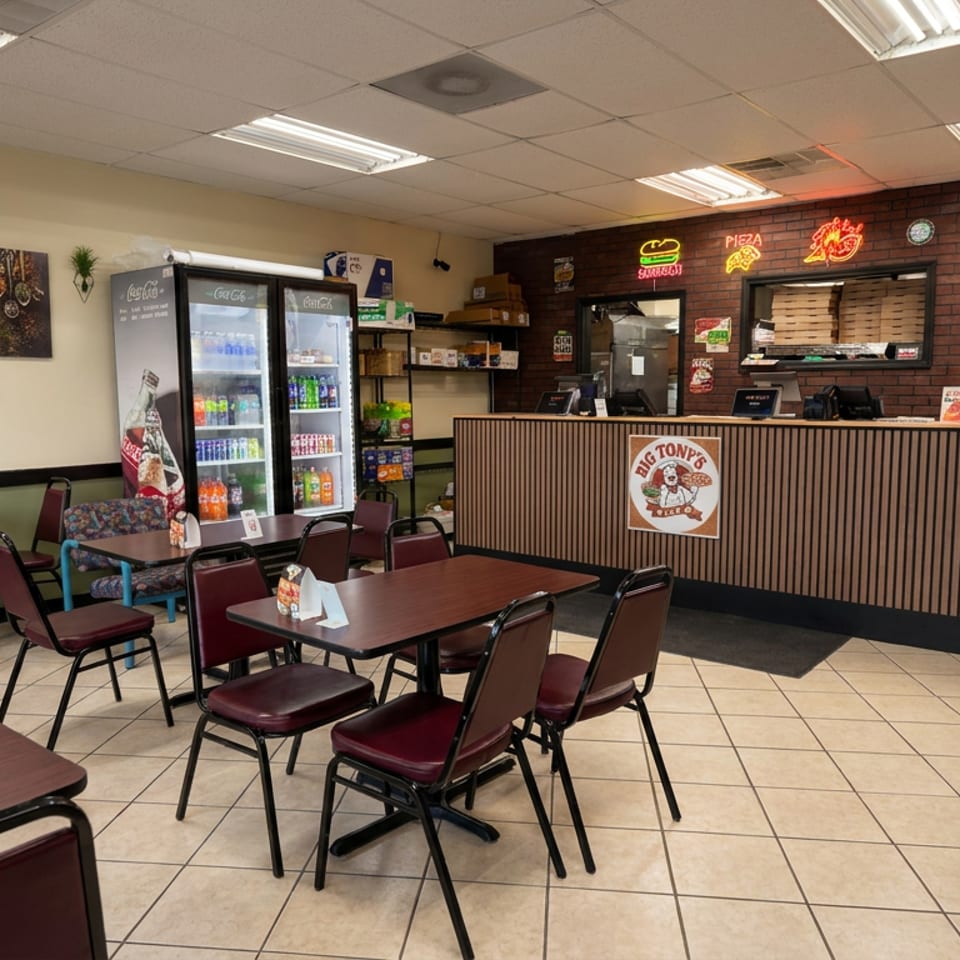 Big Tony’s Pizza dining area with tables, chairs, beverage coolers, food counter, neon signs, and brick accent wall.