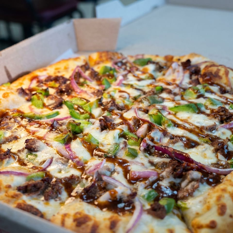 Cheesy pizza topped with ground meat, green peppers, red onions, mushrooms, and barbecue sauce in an open cardboard box.