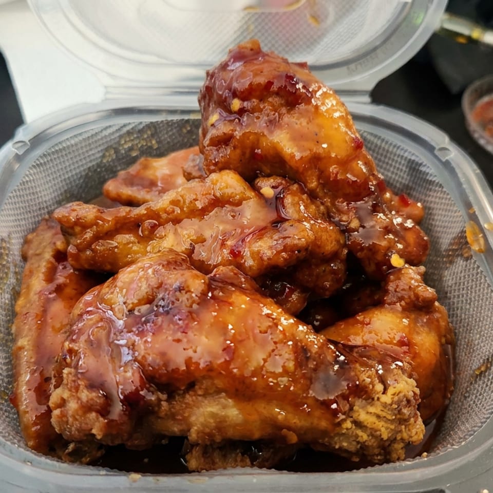 Close-up of glazed fried chicken pieces piled in a clear takeout container, with dipping sauce nearby.