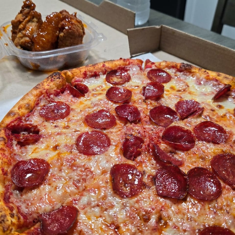 Pepperoni pizza in an open cardboard box with a container of glazed chicken wings behind it on a table.